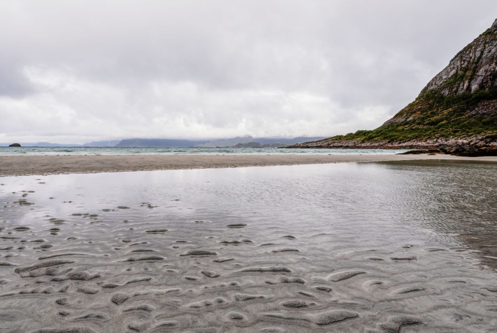 Vindfull sandstrand med fjord, fjell og svaberg under grå himmel