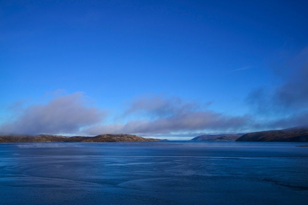 Rolig fjord med blått vann, lave fjell og skyer over horisonten